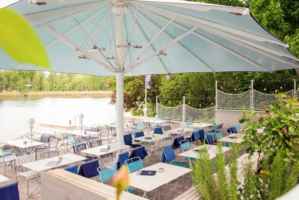 Restaurant mit Sitzgelegenheiten im Freien an einem See, mit blauen und weißen Tischen und Stühlen unter einem großen hellblauen Sonnenschirm, umgeben von Grünpflanzen und Bäumen.