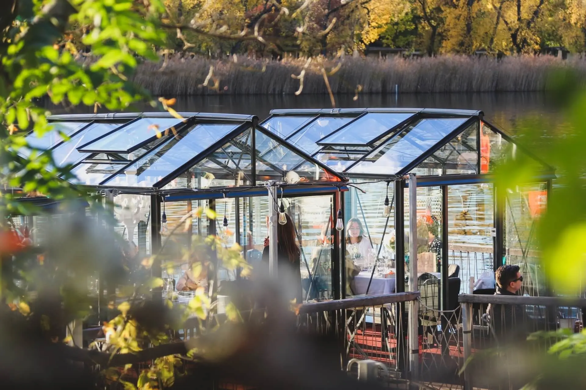 Die gläsernen Speisehallen stehen am Wasser, umgeben von Grün und herbstlichen Bäumen, und die Gäste sitzen drinnen und genießen ihre Mahlzeiten in einer gemütlichen, geschlossenen Umgebung.