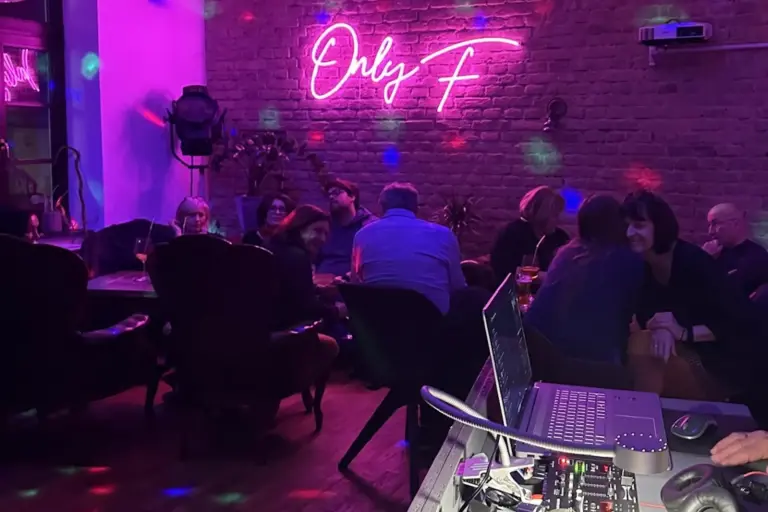 Das F Menschen sitzen und unterhalten sich in einer schwach beleuchteten Bar mit violetter Beleuchtung. Auf einem Neonschild an einer freiliegenden Backsteinmauer steht "Only F". Im Vordergrund ist eine Tonanlage zu sehen, und bunte Lichtpunkte zieren die Szene.
