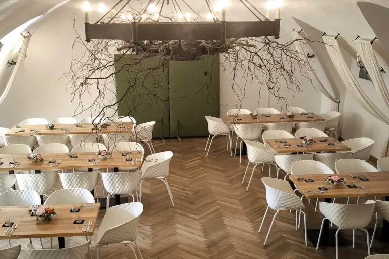 Freiherr Ein moderner Speisesaal mit Holztischen und weißen Stühlen, die für eine Mahlzeit gedeckt sind. Ein großer Kronleuchter mit kahlen Ästen hängt von der Decke über den Tischen. Der Boden hat ein Fischgrätenmuster.