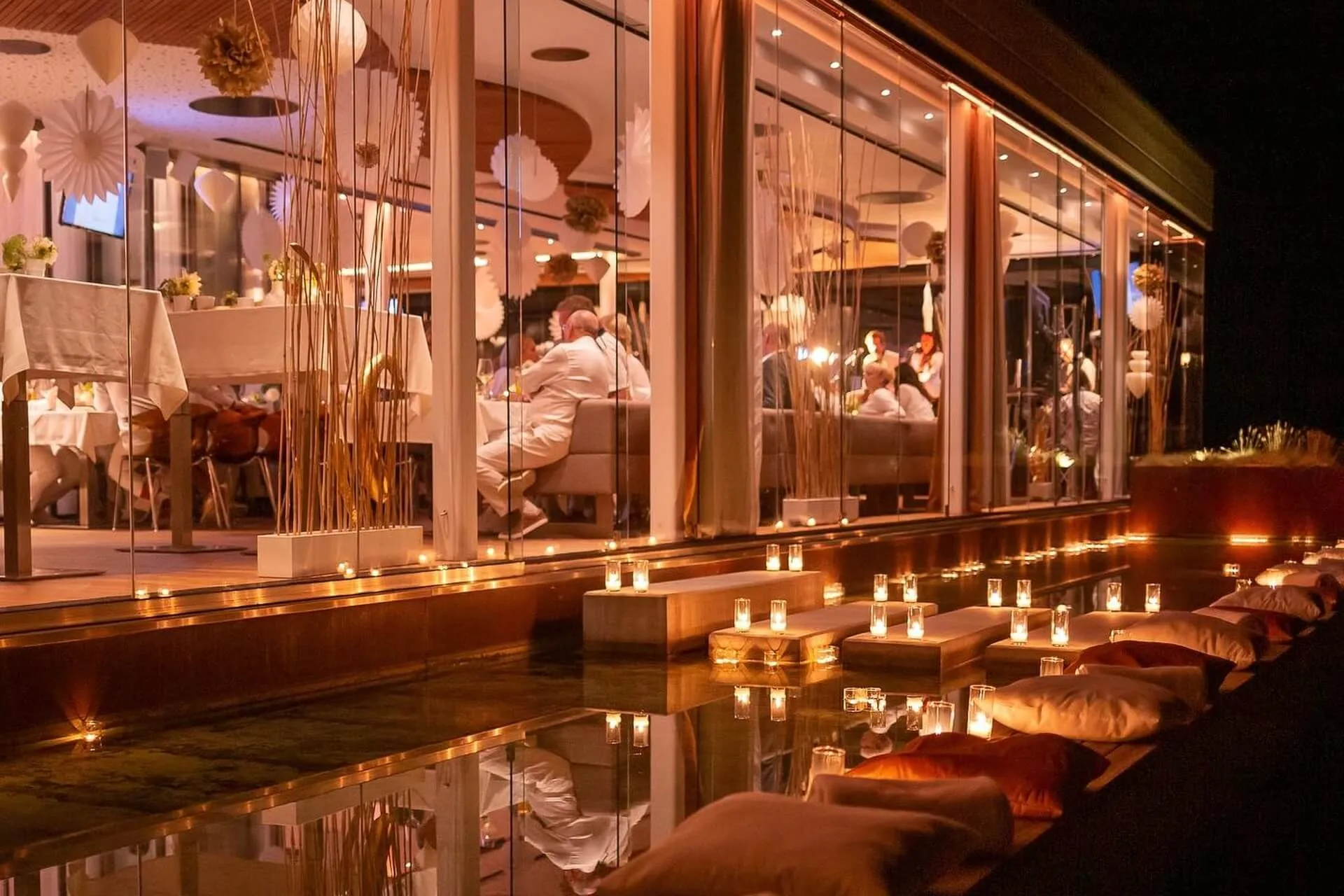 Ein warm beleuchtetes Restaurant mit großen Fenstern zeigt die Gäste beim Essen im Inneren. Draußen säumen Kerzen den Rand des Wassers mit Sitzkissen, die nachts ein gemütliches, romantisches Ambiente schaffen.