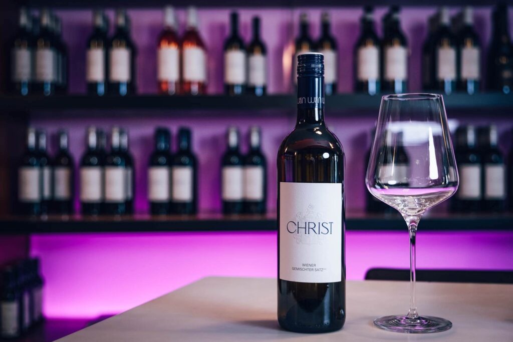 Eine Flasche CHRIST-Wein und ein leeres Weinglas auf einem Tisch vor einem Regal mit verschiedenen Weinflaschen und einem lila beleuchteten Hintergrund - eine TOP Location für Weinliebhaber.