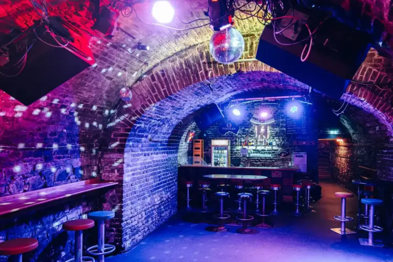 Eine schummrig beleuchtete Bar mit gewölbten Ziegeldecken, bunten Scheinwerfern, einer Discokugel, hohen Hockern und einem Tresen im hinteren Bereich. Die Atmosphäre ist lebendig und hat das Gefühl eines Nachtclubs im Untergrund.