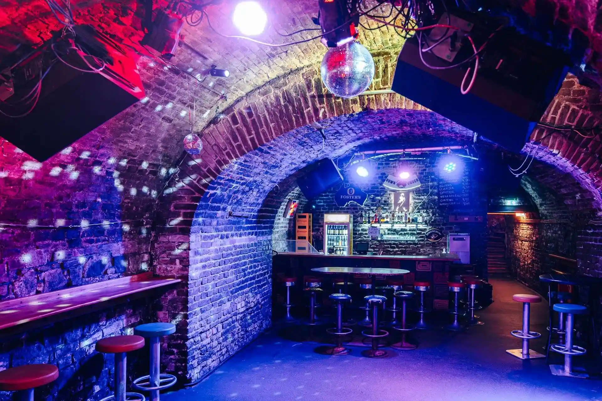 Eine schummrig beleuchtete Bar mit gewölbten Ziegeldecken, bunten Scheinwerfern, einer Discokugel, hohen Hockern und einem Tresen im hinteren Bereich. Die Atmosphäre ist lebendig und hat das Gefühl eines Nachtclubs im Untergrund.
