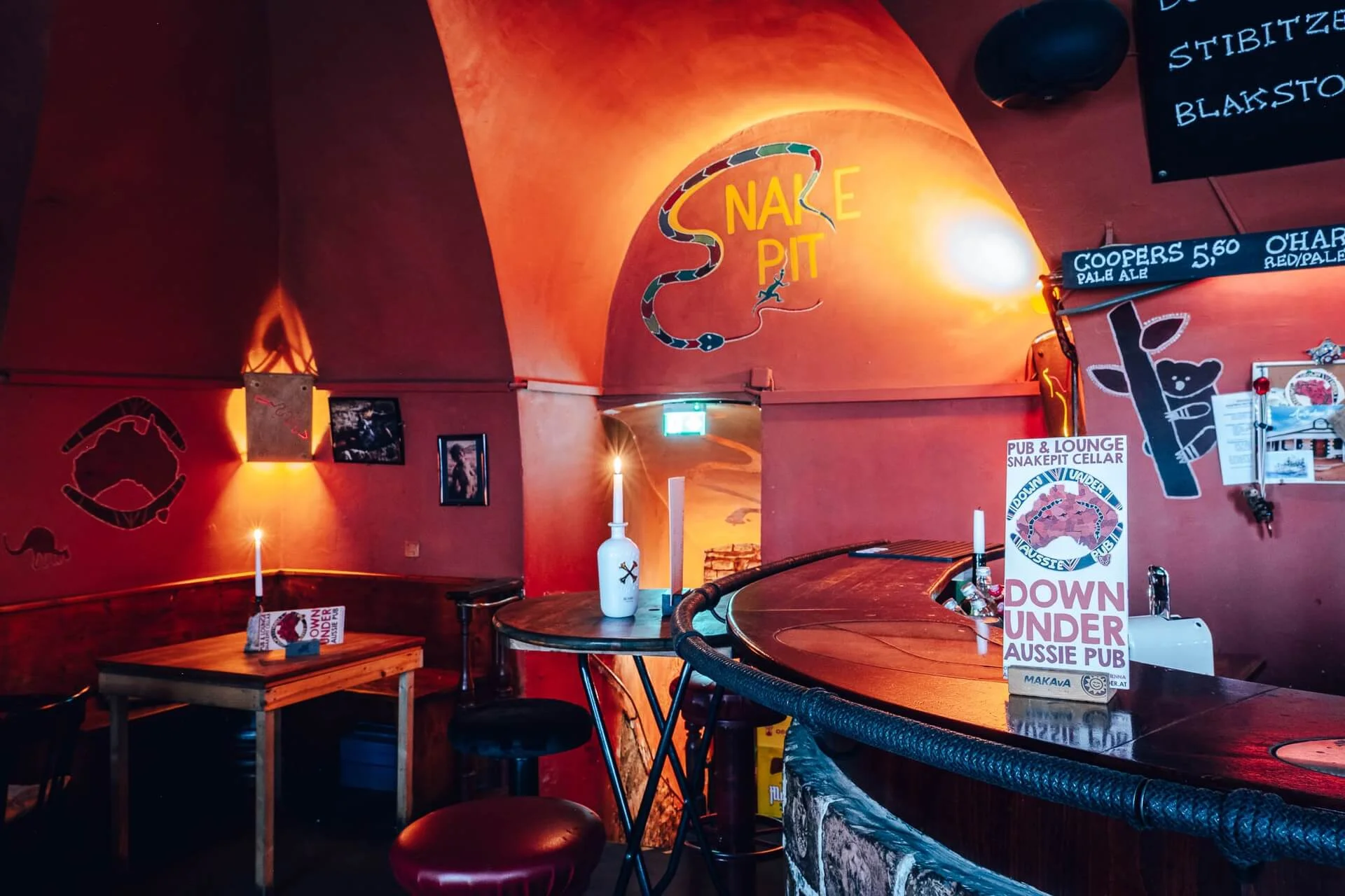 Eine gemütliche Bar mit roten Wänden, kerzenbeleuchteten Tischen und einem geschwungenen Tresen. Ein Schild mit der Aufschrift Snake Pit und einer Schlangengrafik befindet sich an der gewölbten Wand. Plakate, Speisekarten und gedämpftes Licht schaffen eine warme, einladende Atmosphäre.