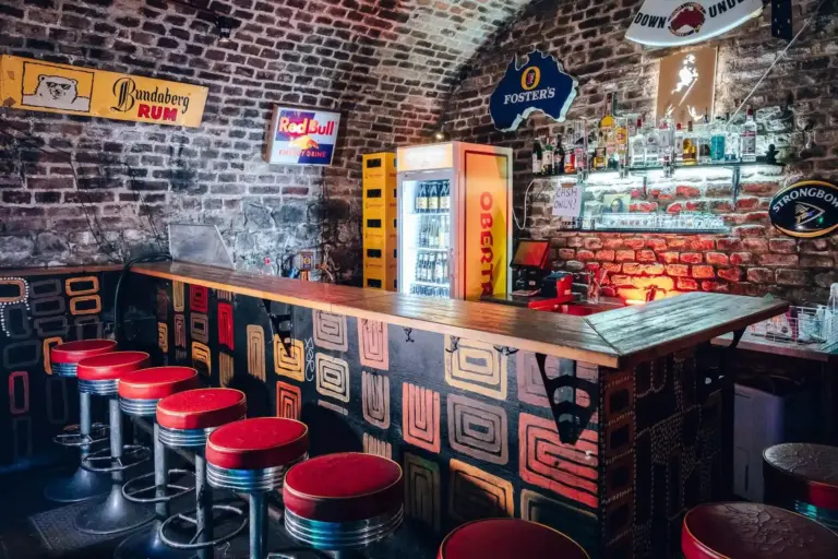 Eine gemütliche Bar mit rot gepolsterten Hockern, einem gemusterten Holztresen und freigelegten Backsteinwänden, die mit alten Schildern verziert sind. Hinter der Theke stehen Kühlschränke mit Getränken und Regale mit Flaschen und Gläsern.