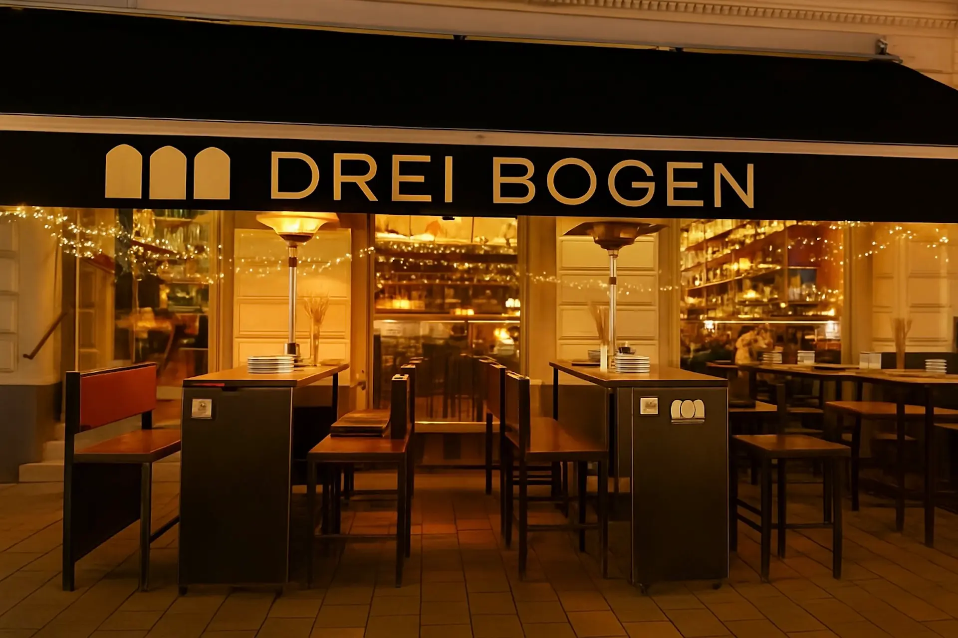 Außenansicht eines Restaurants namens DREI BOGEN mit leeren Tischen und Stühlen auf einer Terrasse, warmer Beleuchtung im Inneren und Lichterketten als Dekoration am Fenster.
