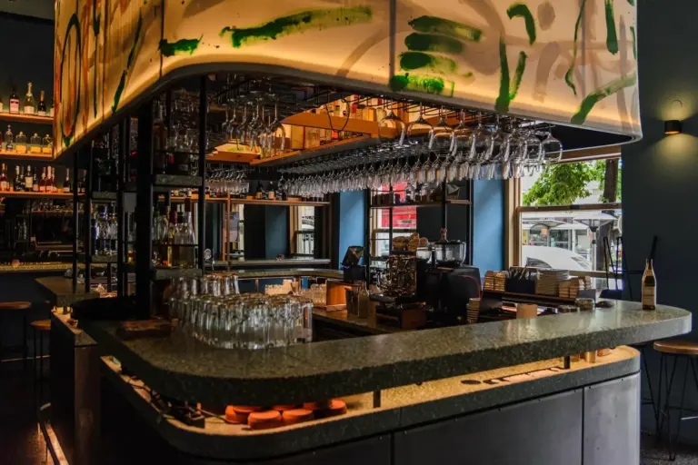 Drei Bögen Eine moderne Bar mit einer grün gesprenkelten Theke, hängenden Weingläsern, Regalen voller Flaschen und großen Fenstern, durch die Tageslicht hereinströmt.
