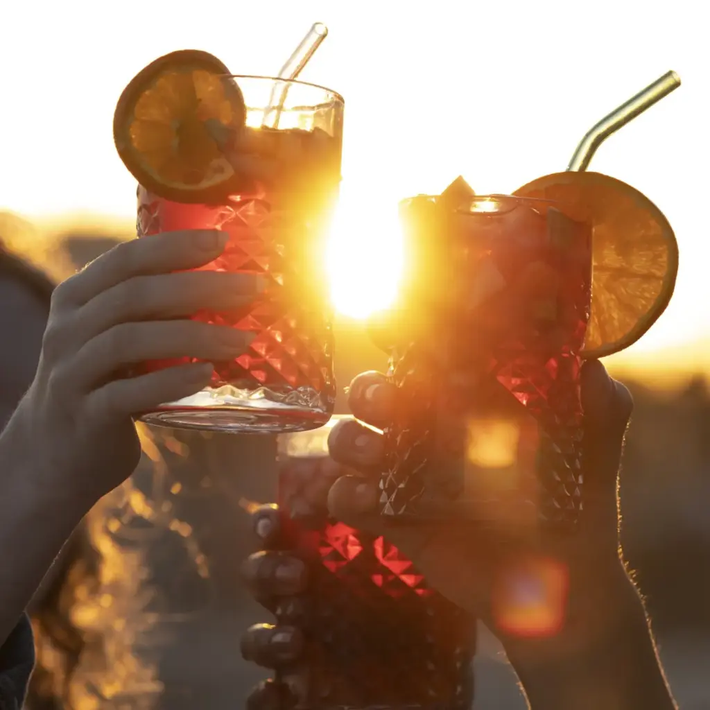Drei Hände erheben Gläser mit einem roten Getränk, das mit Orangenscheiben und Glasstrohhalmen garniert ist, während im Hintergrund die Sonne hell scheint - so wird die fröhliche Stimmung eines Sommerfestes in Wien eingefangen.