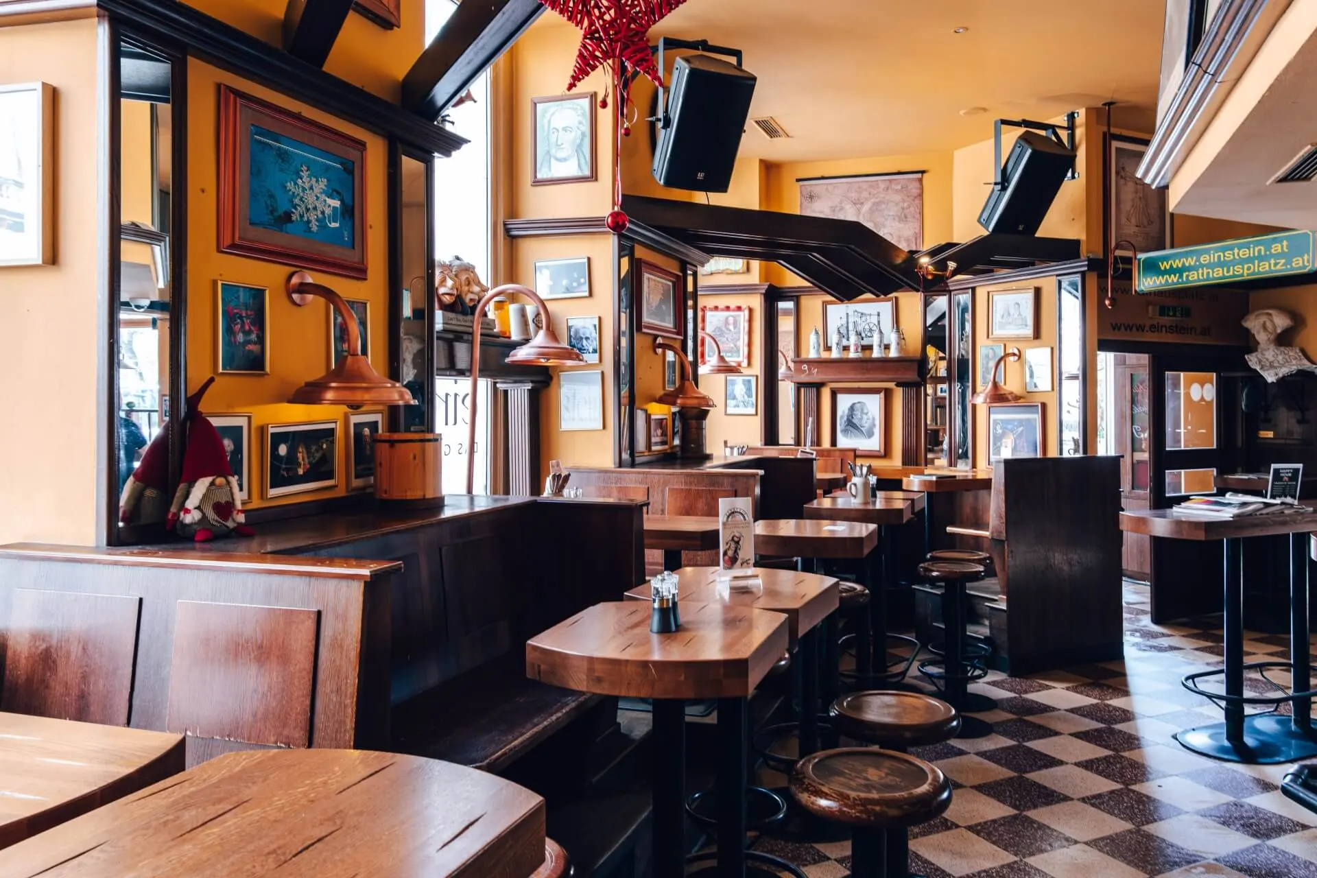 Ein gemütliches, leeres Café mit hölzernen Ständen und Tischen, Hockern, warmen gelben Wänden und gerahmten Bildern. Von der Decke hängen kupferne Leuchten, und durch große Fenster strömt Sonnenlicht herein.