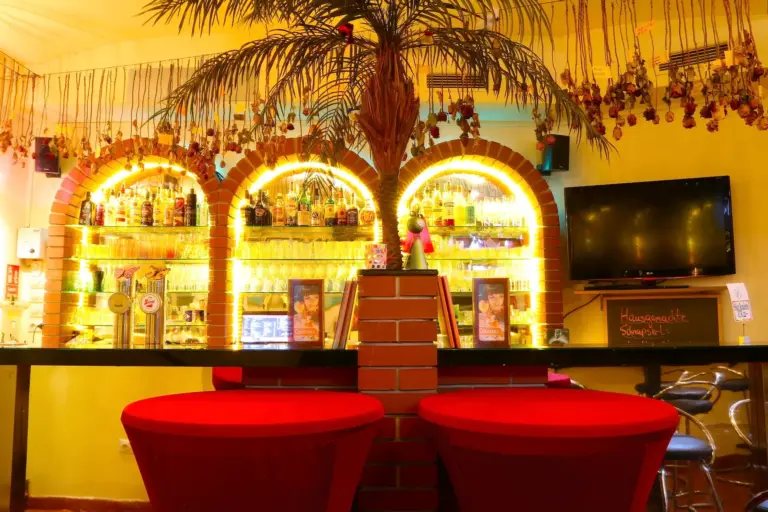 Eine lebhafte Bar mit roten Hockern, einem gemauerten Tresen und einer dekorativen künstlichen Palme. In drei gewölbten Nischen hinter dem Tresen sind Regale mit Flaschen beleuchtet, und rechts an der Wand hängt ein Fernseher.