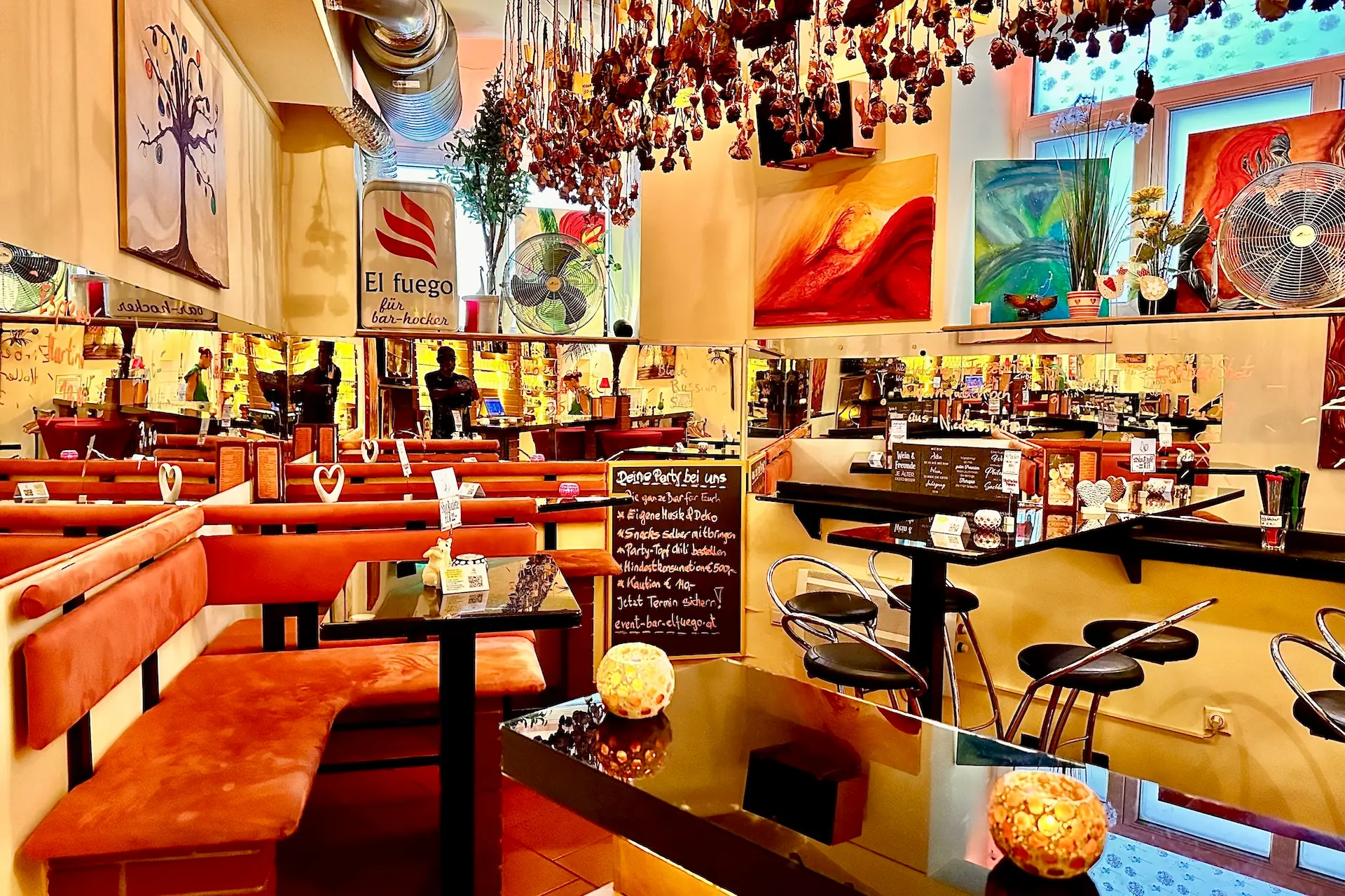 Ein gemütliches, hell beleuchtetes Café mit orangefarbenen, gepolsterten Bänken, Wandkunst, hängenden Dekorationen und einer Speisekarte. An den Wänden befinden sich Spiegel, die die Tische und die warme Beleuchtung reflektieren und eine einladende Atmosphäre schaffen.