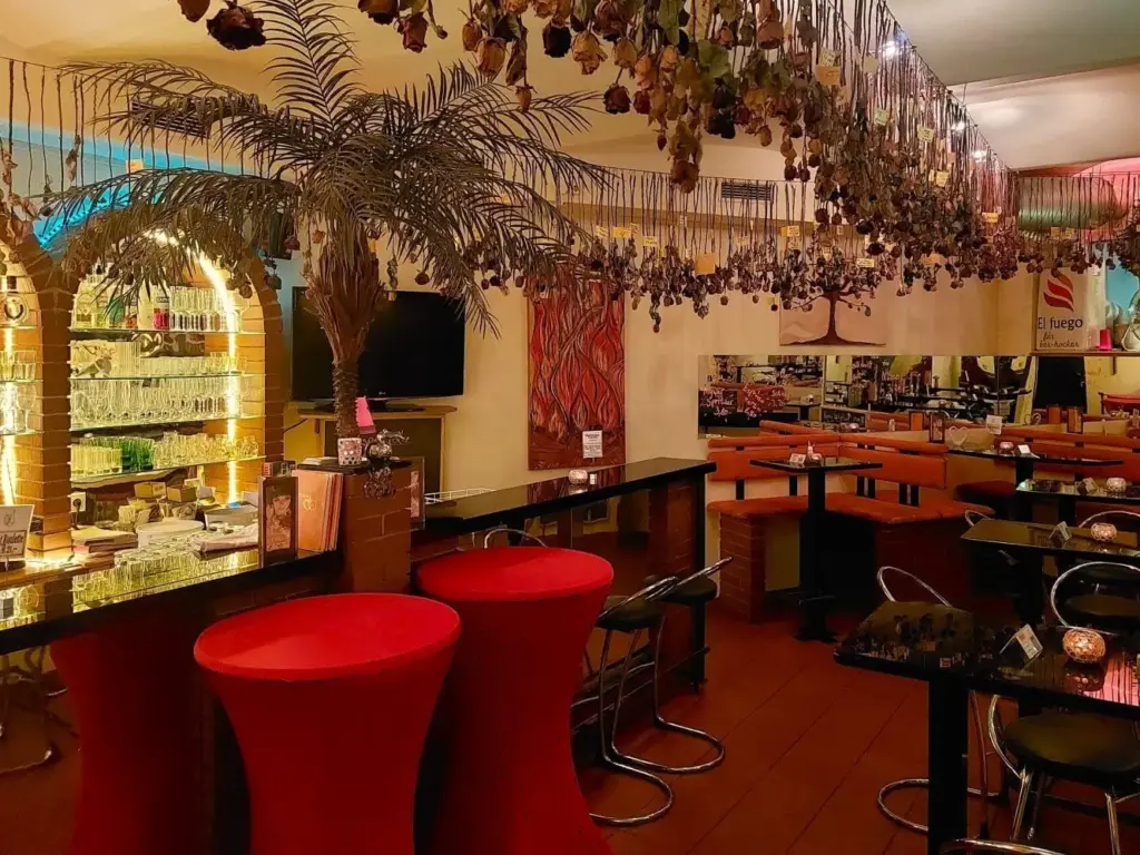Das El fuego Wien ist ein gemütliches Restaurant mit roten Barhockern, einem Tresen mit Getränken und Tischen mit Stühlen. Getrocknete Pflanzen hängen von der Decke, und die warme Beleuchtung schafft eine einladende Atmosphäre. Ein Fernseher und dekorative Elemente sind zu sehen.