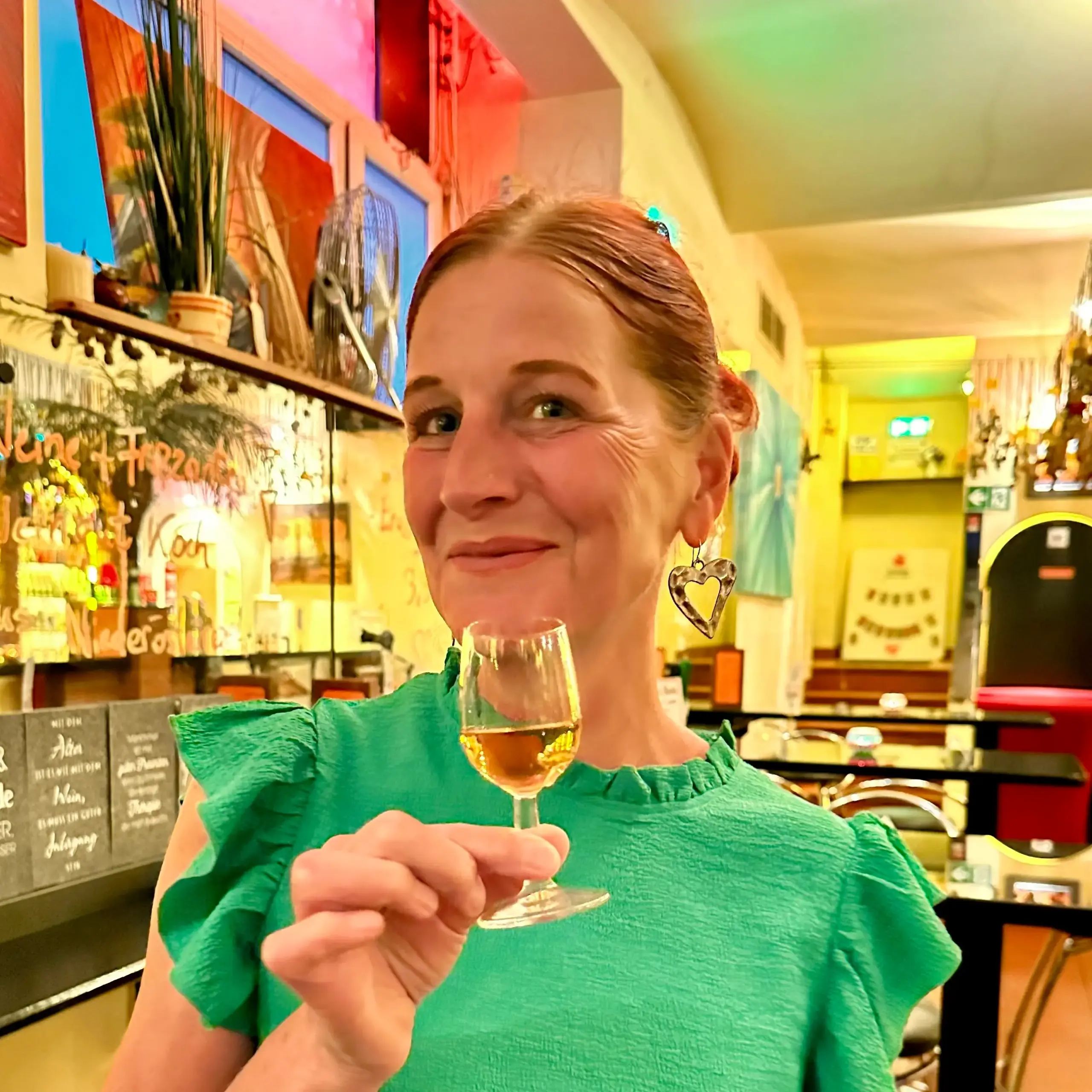 Eine Frau in einem grünen Oberteil lächelt und hält ein Glas mit einem hellen Getränk in einem farbenfrohen, gemütlichen Restaurant namens El Fuego, mit herzförmigen Ohrringen und warmer Umgebungsbeleuchtung.