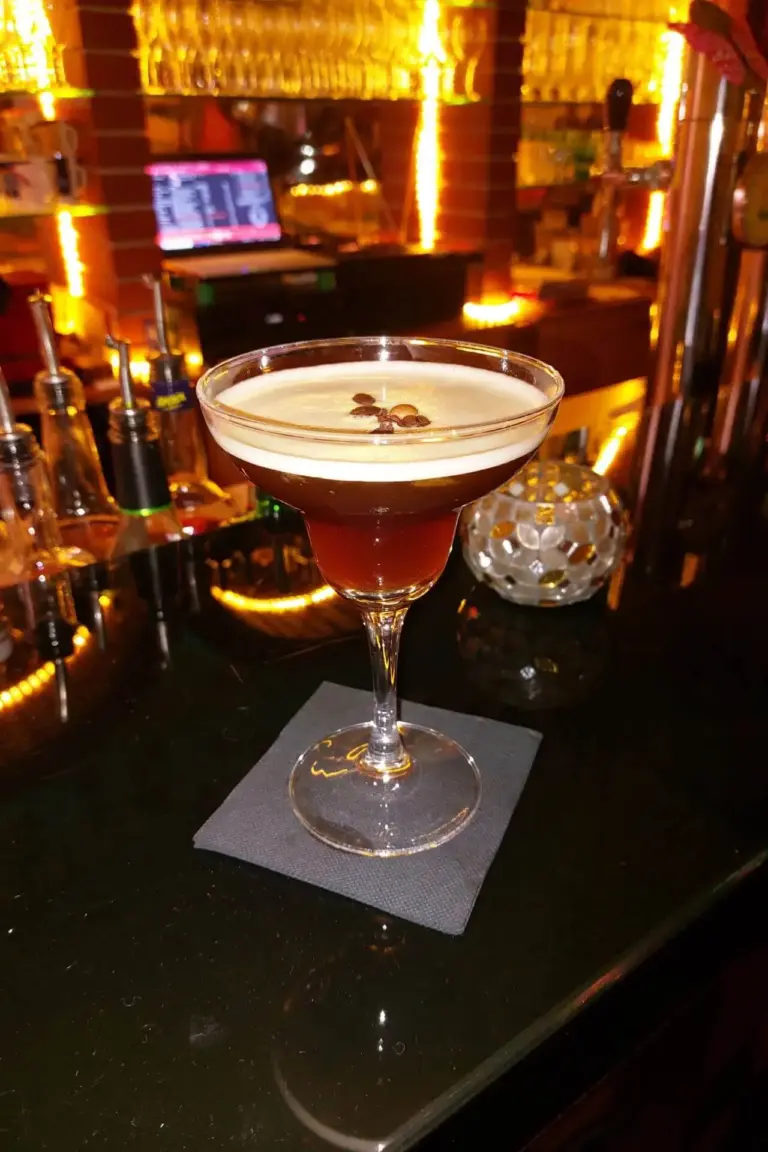 Ein mit einem dunklen Cocktail gefülltes Martiniglas mit Schaum und drei Kaffeebohnen steht auf einer schwarzen Serviette auf einem Bartresen mit warmer, gelber Beleuchtung im Hintergrund.