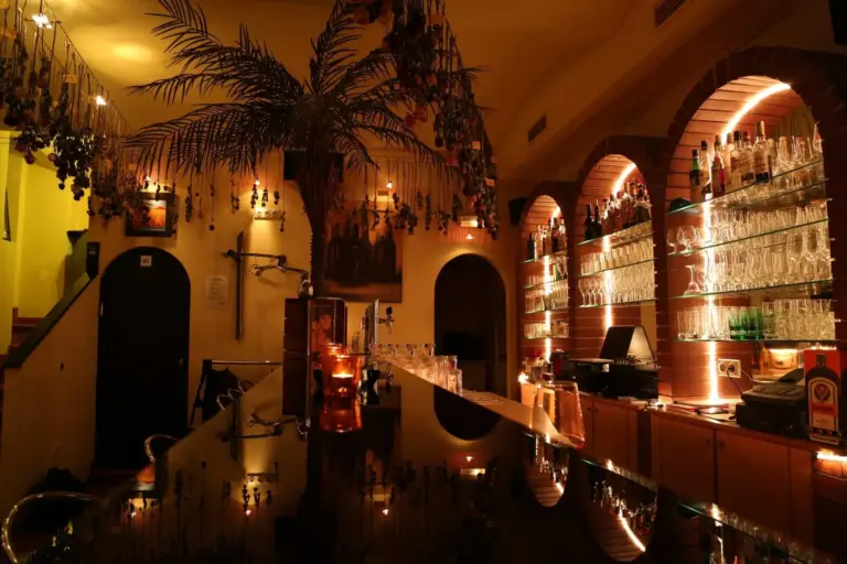 Eine schwach beleuchtete Bar mit gewölbten Regalen, in denen Flaschen und Gläser ausgestellt sind, warme gelbe Beleuchtung, eine Palme in der Mitte und Kerzen schaffen eine gemütliche, einladende Atmosphäre.