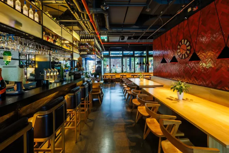 Modernes Bar- und Restaurantinterieur mit einem langen Holztisch, Holzstühlen, einem Bartresen mit Barhockern, hängenden Gläsern, einer rot gefliesten Wand, einer großen Uhr und Pendelleuchten. Durch die großen Fenster an der Vorderseite strömt Sonnenlicht herein.