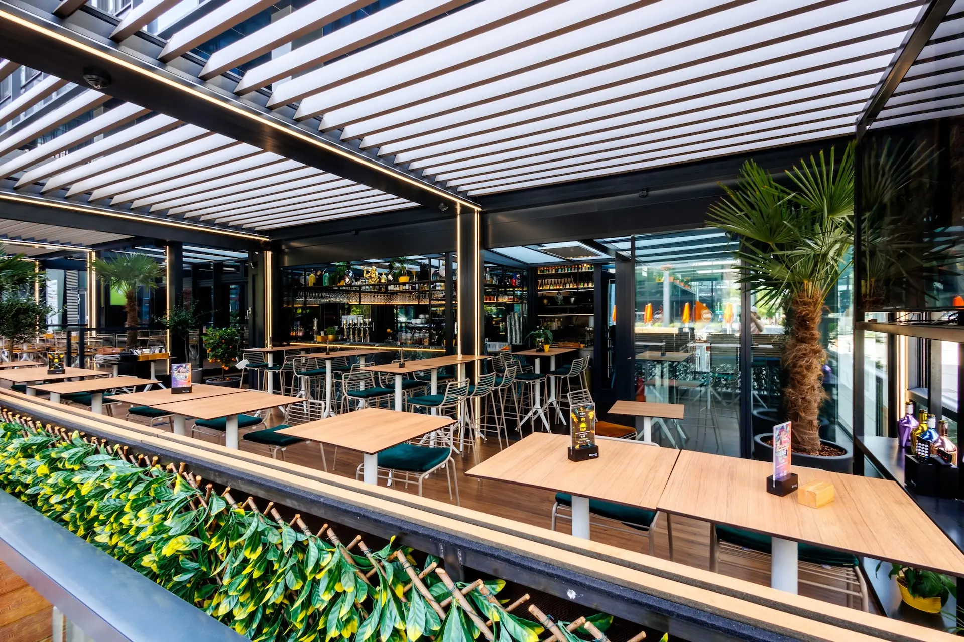 Modernes Außenrestaurant mit Holztischen, grünen Stühlen und getopften Palmen. Das Lamellendach lässt viel Licht herein. Üppige Grünpflanzen säumen den Essbereich, und im Hintergrund ist eine gut gefüllte Bar zu sehen.