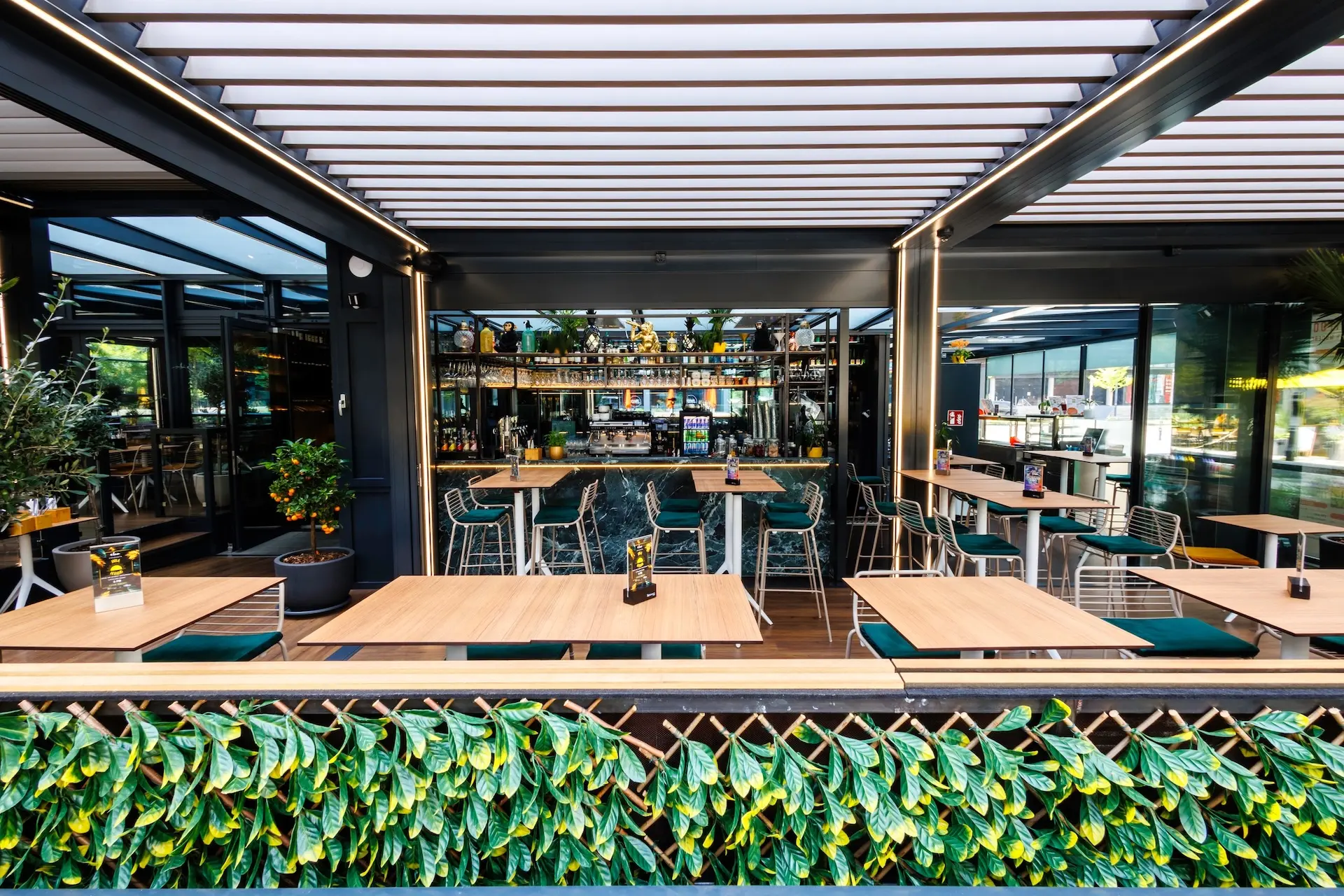 Modernes Außenrestaurant mit Holztischen, grünen Stühlen und Blattschmuck. Im Hintergrund ist der Barbereich mit Flaschen und Gläsern zu sehen, unter einem teilweise überdachten Dach mit viel natürlichem Licht.