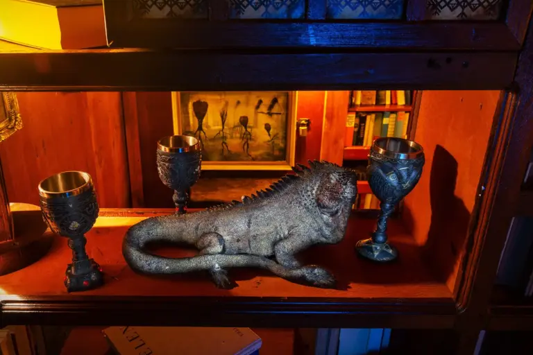 Eine realistische Leguan-Skulptur steht auf einem Holzregal zwischen drei verschnörkelten Kelchen, während im Hintergrund ein warm beleuchteter Raum und ein surreales Gemälde zu sehen sind.