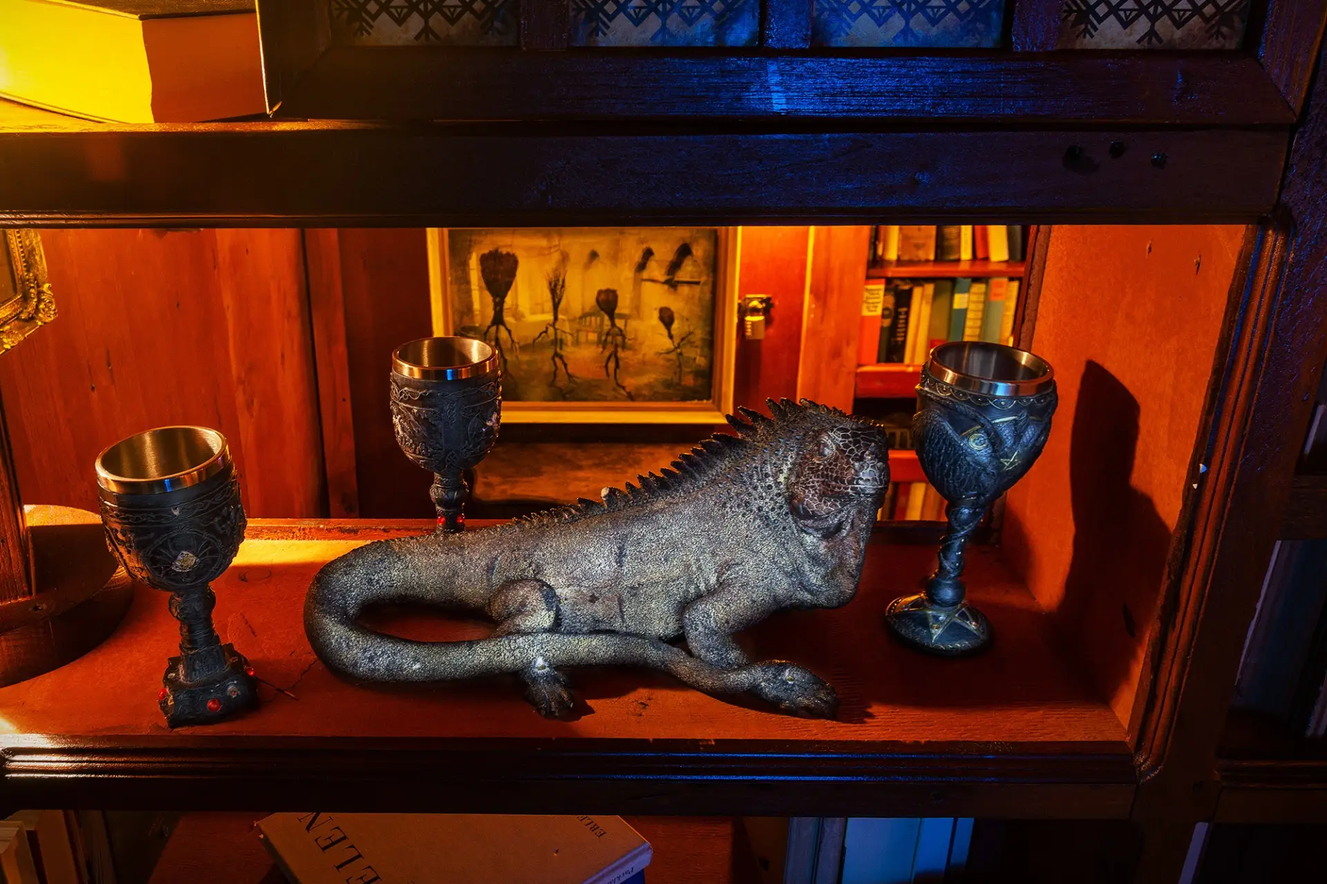 Eine realistische Leguan-Skulptur steht auf einem Holzregal zwischen drei verschnörkelten Kelchen, während im Hintergrund ein warm beleuchteter Raum und ein surreales Gemälde zu sehen sind.