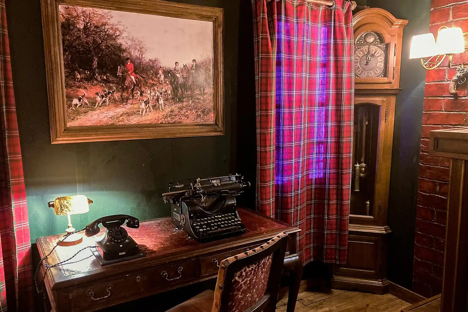 EscapeGame Salzburg Ein altes Arbeitszimmer mit einem hölzernen Schreibtisch, auf dem ein Telefon mit Drehscheibe, eine alte Schreibmaschine und eine Lampe stehen. Hinter dem Schreibtisch befinden sich ein gerahmtes Gemälde, rotkarierte Vorhänge und eine große antike Standuhr.