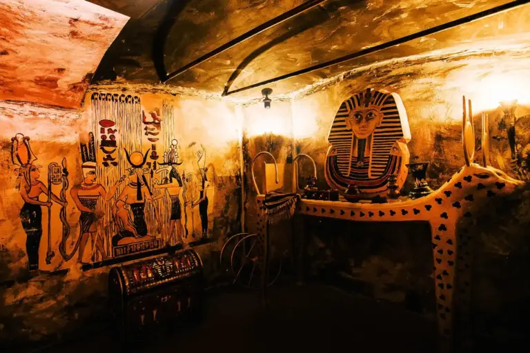 EscapeGame Innsbruck Ein schwach beleuchteter Raum, dekoriert wie ein altägyptisches Grab, mit Wandgemälden ägyptischer Figuren, einer goldenen Maske des Pharaos auf einem Tisch und verschiedenen Schätzen - perfekt für #EscapeGameInnsbruck-Fans, die #Spannung und knifflige Rätsel lieben.