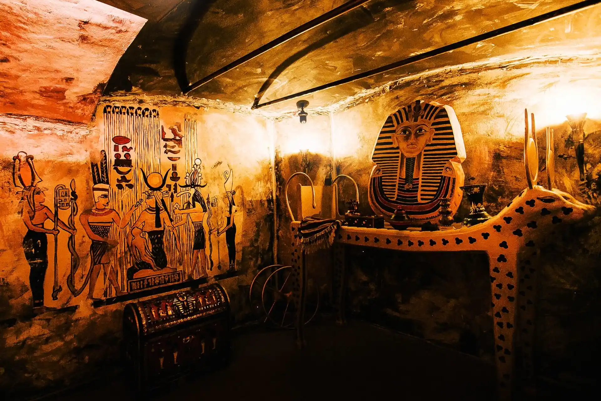 EscapeGame Innsbruck Ein schwach beleuchteter Raum, dekoriert wie ein altägyptisches Grab, mit Wandgemälden ägyptischer Figuren, einer goldenen Maske des Pharaos auf einem Tisch und verschiedenen Schätzen - perfekt für #EscapeGameInnsbruck-Fans, die #Spannung und knifflige Rätsel lieben.