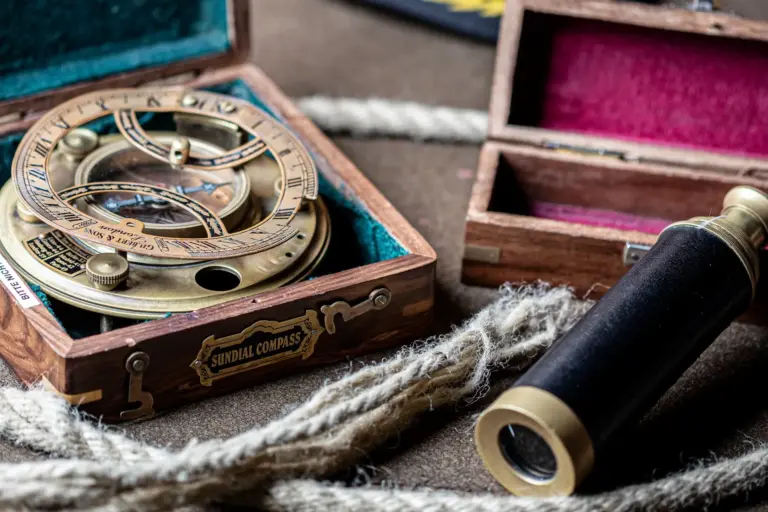 Ein Sonnenuhrkompass aus Messing in einer Holzkiste liegt neben einem alten Fernrohr aus Messing und Leder, beide auf einer strukturierten Oberfläche mit einem dicken Seil in der Nähe - die perfekten Requisiten für ein #EscapeGameInnsbruck voller Spannung und cleverer Raetsel.