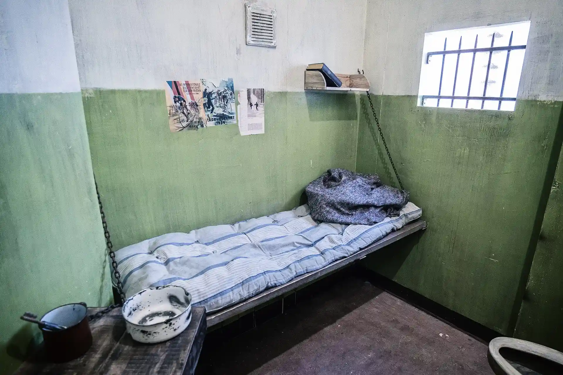 Eine kleine Gefängniszelle mit grünen und weißen Wänden, einem einzelnen Metallbett mit einer dünnen Matratze und einer Decke, einem Regal mit Büchern, einem vergitterten Fenster und einem Tisch mit einer Schüssel und einer Tasse. Über dem Bett sind Plakate an die Wand geklebt.
