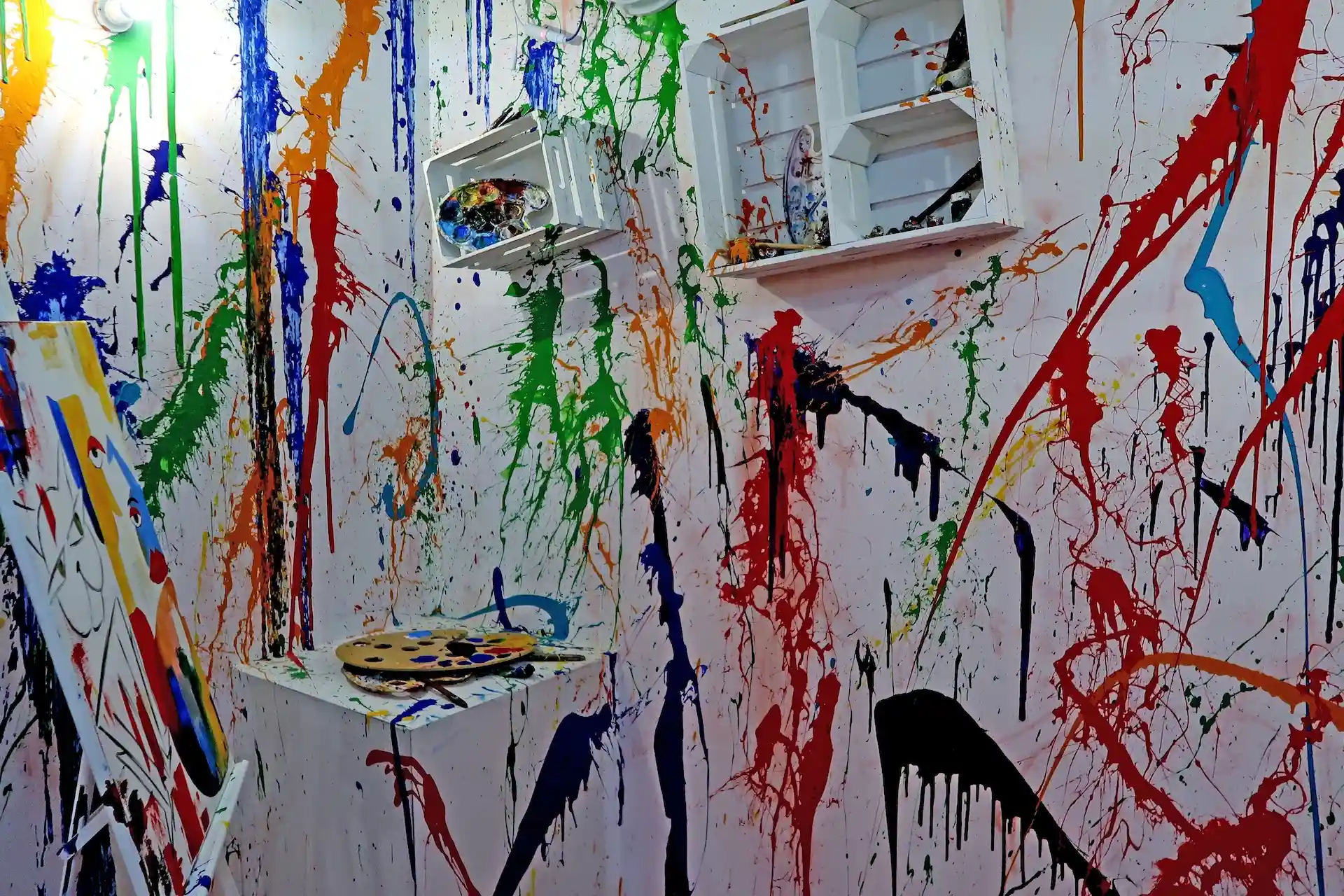 Ein weißer Raum mit Regalen, einer Palette und einer Leinwand, die mit bunten Farbspritzern und -tropfen bedeckt sind und eine chaotische, abstrakte Kunstszene ergeben.