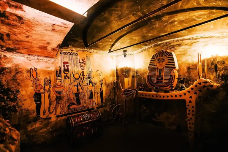 EscapeGame Wien Ein schwach beleuchteter Raum mit ägyptischen Wandmalereien, einer Pharaonenmaske auf einem geschmückten Tisch, einer verzierten Truhe und antiken Symbolen an den Wänden, die eine gruftartige Atmosphäre schaffen.