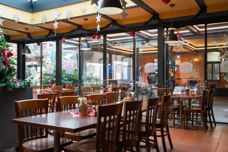 Ein gemütliches Restaurant mit weihnachtlich geschmückten Holztischen und -stühlen, mit Ornamenten, Girlanden und einem Weihnachtsbaum. Große Fenster geben den Blick frei auf festliche Dekorationen und natürliches Licht erfüllt den Raum.