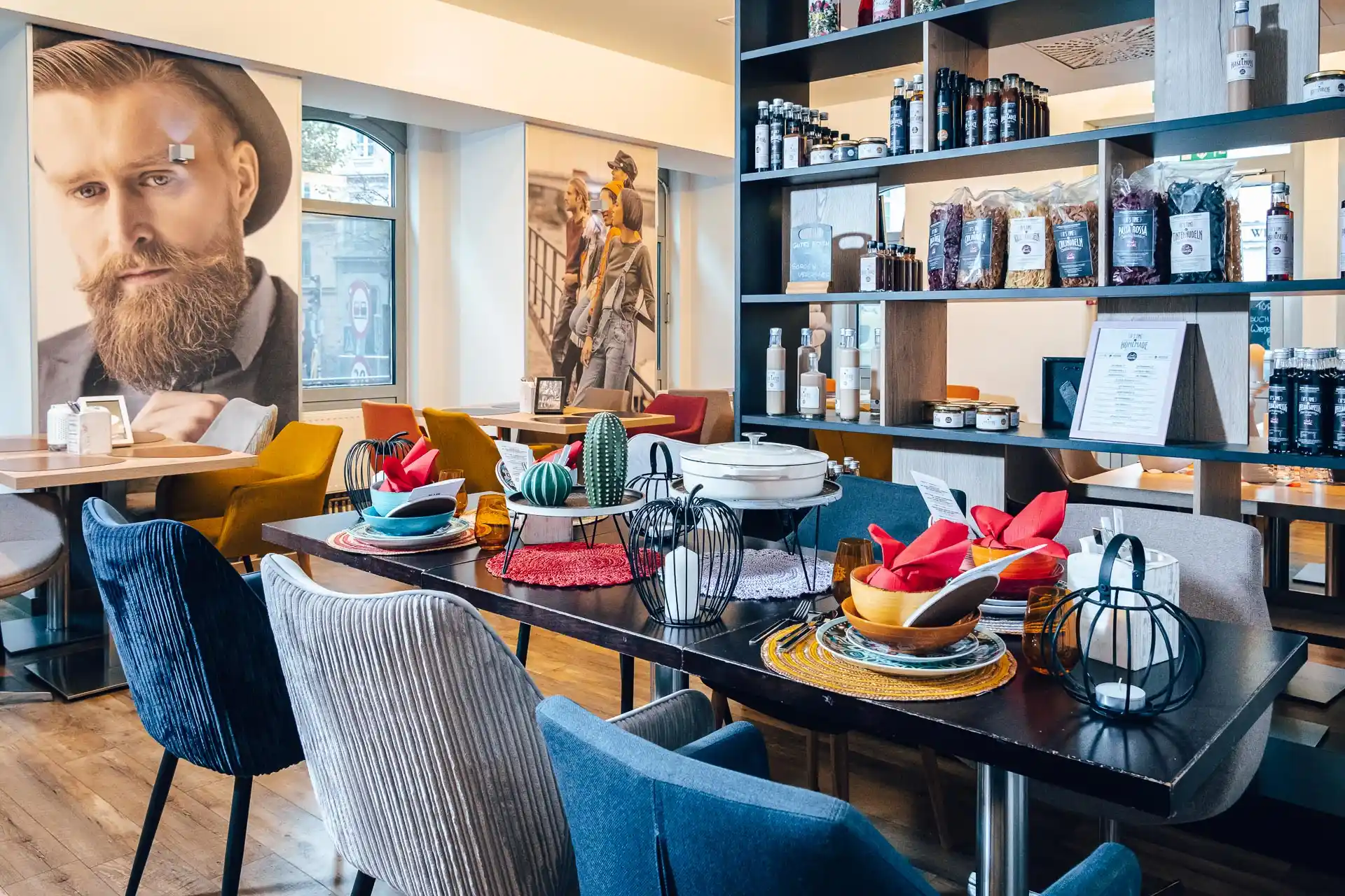 Ein modernes Café mit bunten Stühlen, einem dunklen Tisch mit Tellern, Schalen und roten Servietten, dekorativen Kakteen und Regalen mit Lebensmitteln. Große Porträts schmücken die Wände. Der Raum ist von natürlichem Licht durchflutet.
