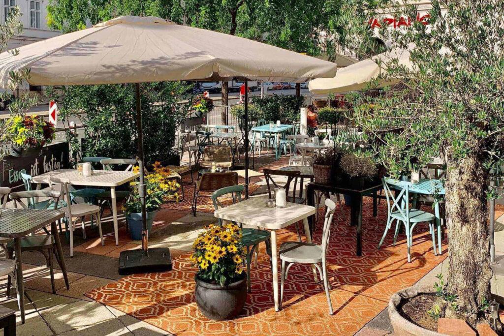 Schanigarten-Café im Freien mit weißen und blauen Stühlen, Tischen, großen Sonnenschirmen, Blumen und Grünzeug auf einem gemusterten Teppich. Das Sonnenlicht fällt durch die Bäume auf den gemütlichen Sitzbereich neben einer belebten Straße.