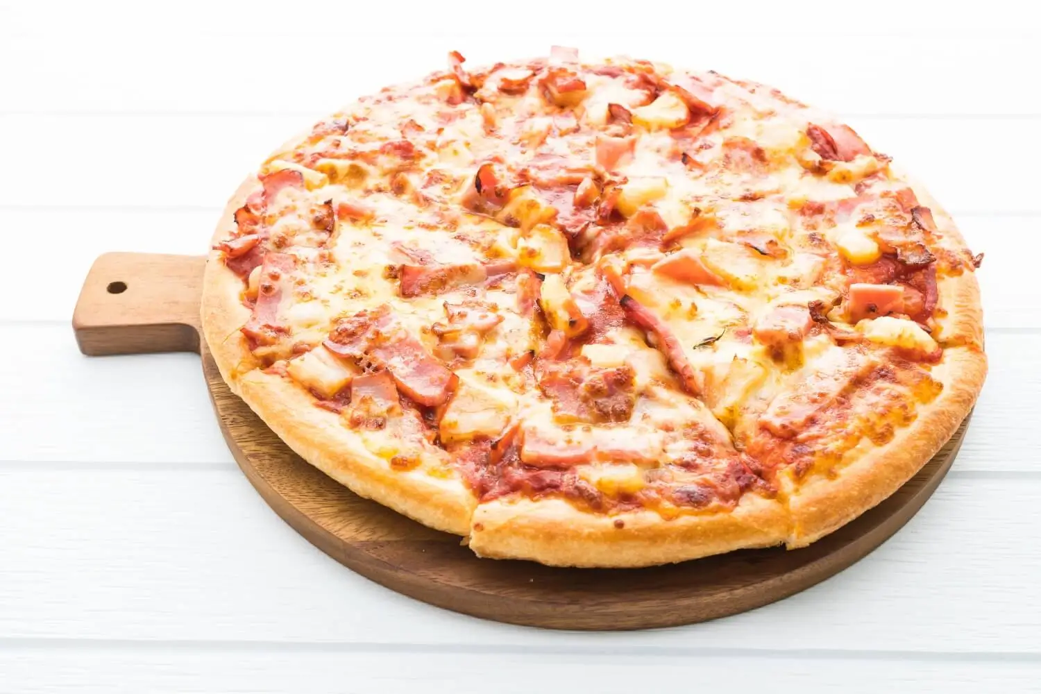 Eine ganze Schinken-Käse-Pizza mit goldener Kruste, aufgeschnitten und serviert auf einem runden Holzpizzabrett vor weißem Hintergrund - ein unwiderstehliches Faschingsrezept für jeden festlichen Anlass.