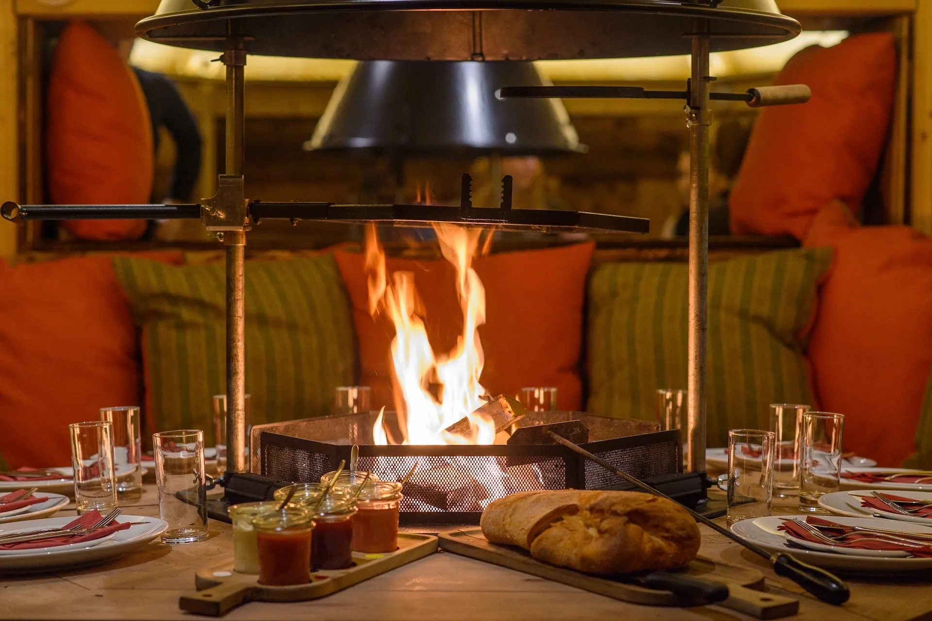 Ein gemütliches Esszimmer-Set mit Brot, Aufstrichen und Gläsern umgibt ein offenes Kaminfeuer in der Mitte, im Hintergrund liegen Kissen, was eine warme, einladende Atmosphäre schafft.