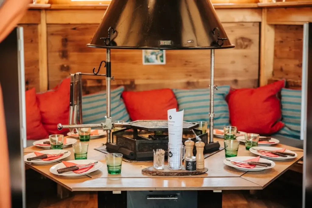 Ein gemütlicher Essbereich mit einem runden Tisch für sechs Personen, gedeckt mit grünen Gläsern, Servietten, Besteck und Gewürzen. In der Mitte steht ein großer Grill, umgeben von roten und orangefarbenen Kissen auf einer Holzbank.