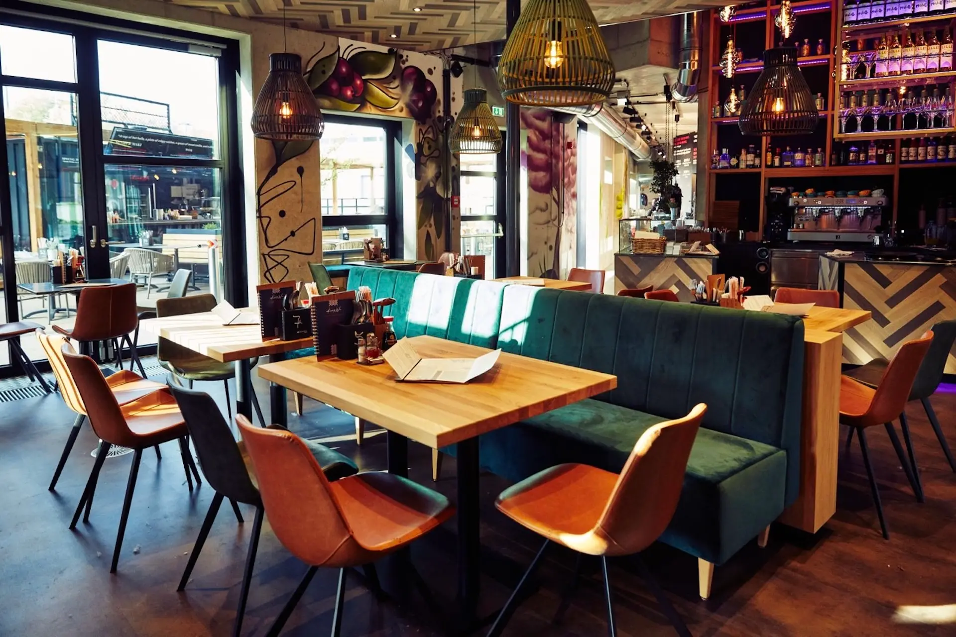 Modernes Restaurantinterieur mit Holztischen, gepolsterten Sitznischen in Teal und braunen Stühlen. Große Fenster lassen natürliches Licht herein. Von der Decke hängen Hängelampen, und im Hintergrund ist der Barbereich zu sehen.