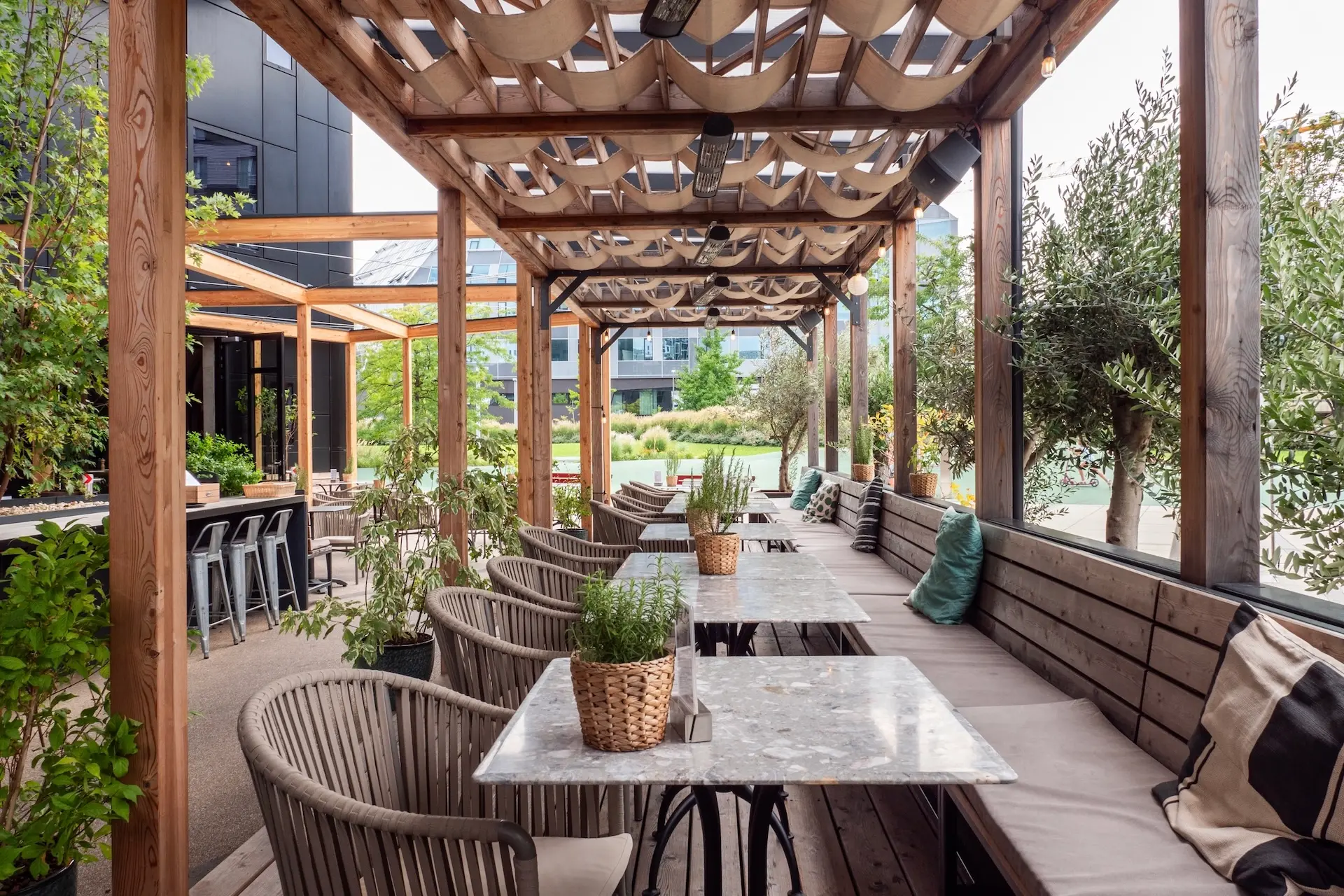 Restaurantterrasse im Freien mit Holzpergola, Marmortischen, Korbstühlen und gepolsterten Bänken. Topfpflanzen schmücken die Tische und natürliches Grün umgibt den Sitzbereich. Im Hintergrund sind moderne Gebäude zu sehen.