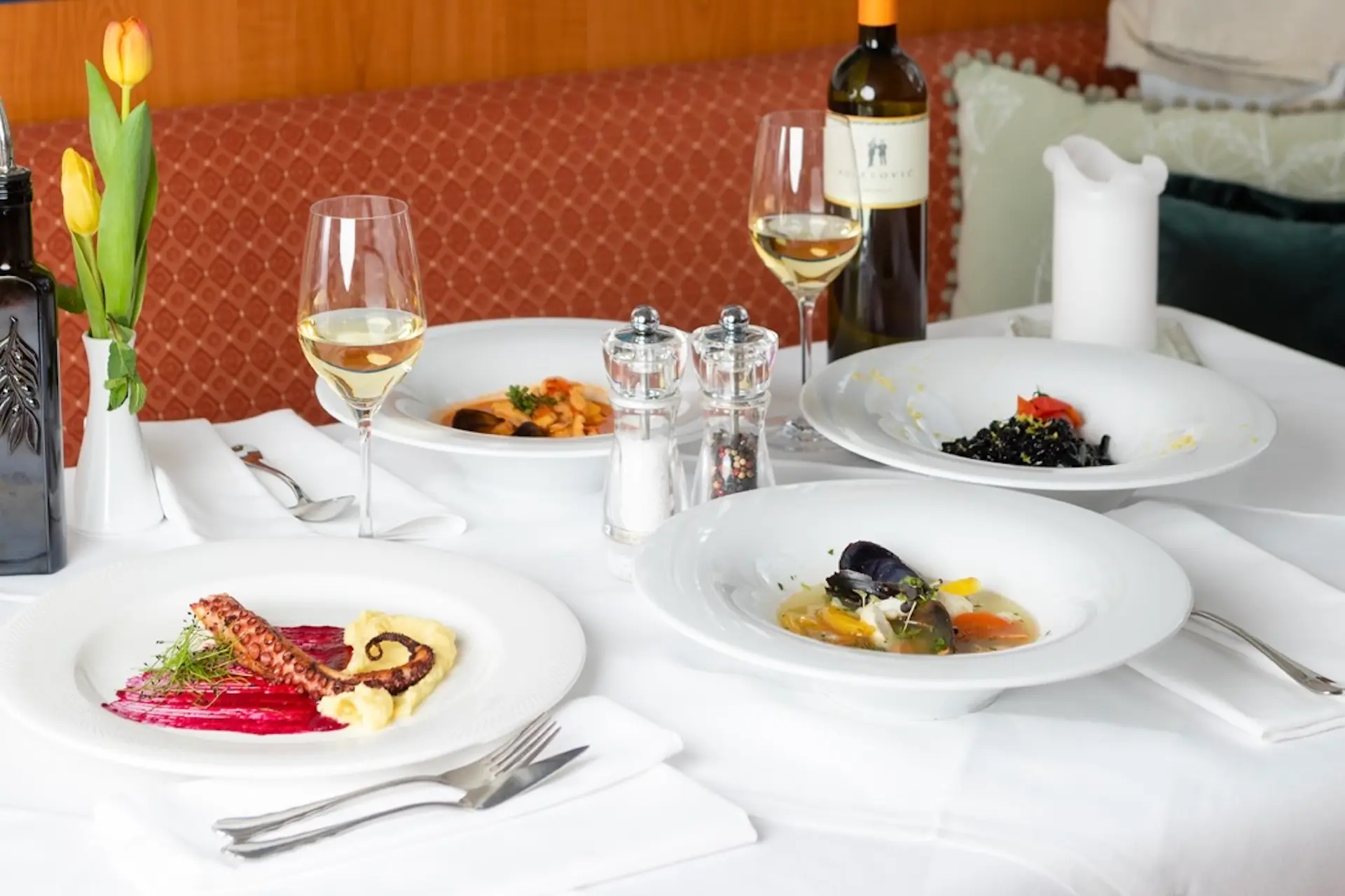 Ein mit weißem Tischtuch gedeckter Esstisch mit elegantem Meeresfrüchtegeschirr, zwei Weißweingläsern, einer Flasche Wein, Salz- und Pfefferstreuern und einem Strauß gelber Tulpen.