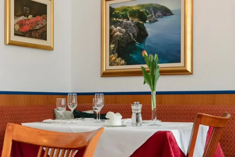 Ein ordentlich gedeckter Restauranttisch mit weißer Tischdecke, rotem Akzenttuch, Gläsern, Besteck, einer Tulpe in einer Vase, Salz- und Pfefferstreuern und einem gerahmten Kunstwerk mit einer Küstenlandschaft an der Wand dahinter.