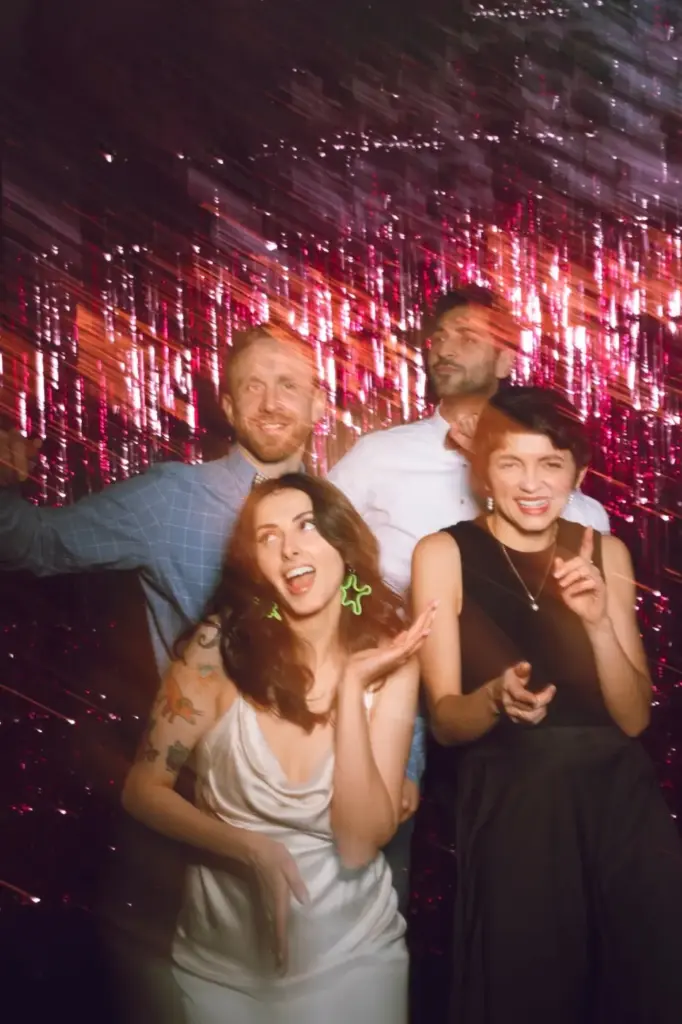 Vier Personen posieren und lächeln spielerisch auf einer After-Wedding-Party, umgeben von glitzernder rosa und lila Lametta-Dekoration, die eine festliche und lebhafte Atmosphäre schafft.