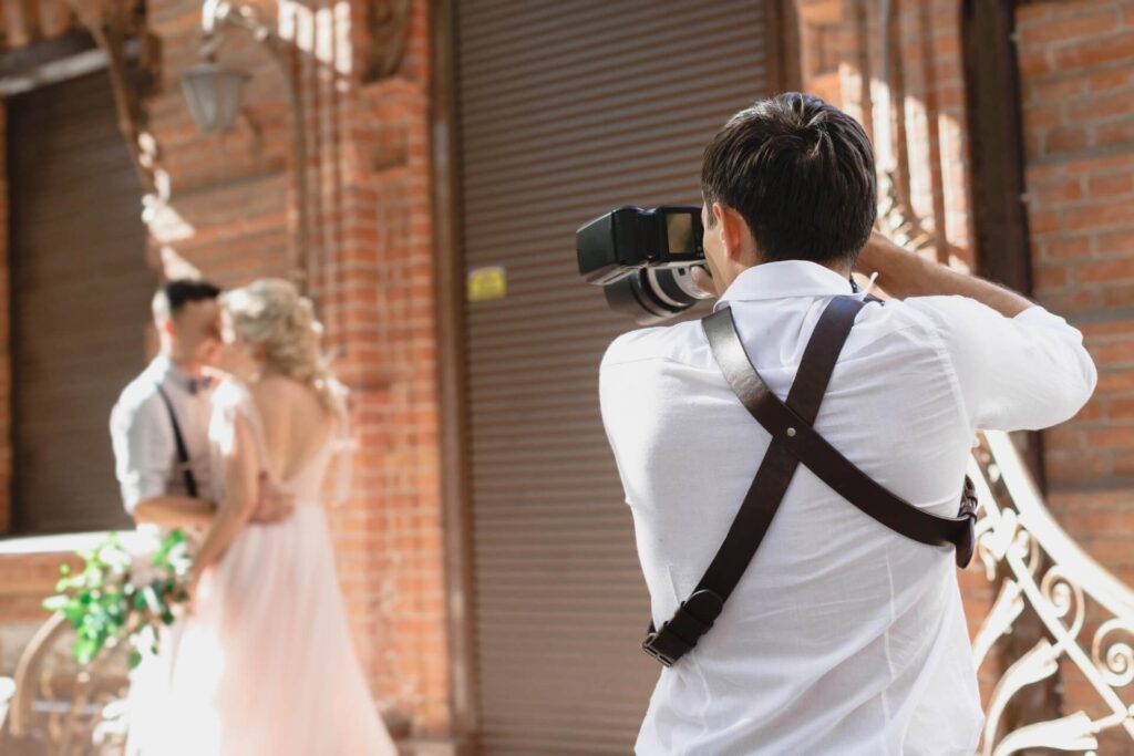 Ein Fotograf für Hochzeit in einem weißen Hemd fängt ein Bild eines Paares ein, das sich vor einem Backsteingebäude umarmt. Das Paar ist leicht unscharf, während der Fotograf im Fokus ist und diesen romantischen Hochzeitsmoment festhält.