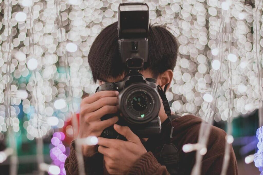 Eine Person, die eine Kamera mit Blitzlicht in der Hand hält, macht ein Foto auf einer lebhaften Veranstaltung; die verschwommenen, leuchtenden Lichterketten im Hintergrund erzeugen einen Bokeh-Effekt, die ideale Inspiration, wenn Sie eine Partylocation mieten möchten.