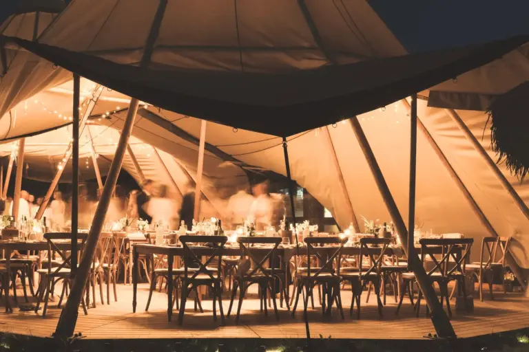 Ein warm beleuchtetes Außenzelt mit dekorierten Tischen und Holzstühlen für ein nächtliches Abendessen. Darüber hängen Lichterketten, und im Inneren bewegen sich verschwommene Figuren, die eine festliche Atmosphäre schaffen.