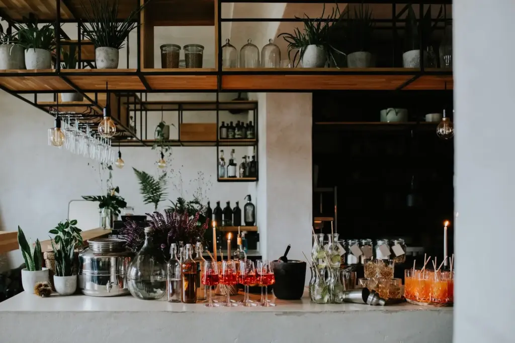 Ein moderner Bartresen mit Gläsern, Flaschen, Pflanzen und mehreren bunten Cocktails. Darüber hängen Regale mit Gläsern und Grünzeug, die eine stilvolle und einladende Atmosphäre schaffen.