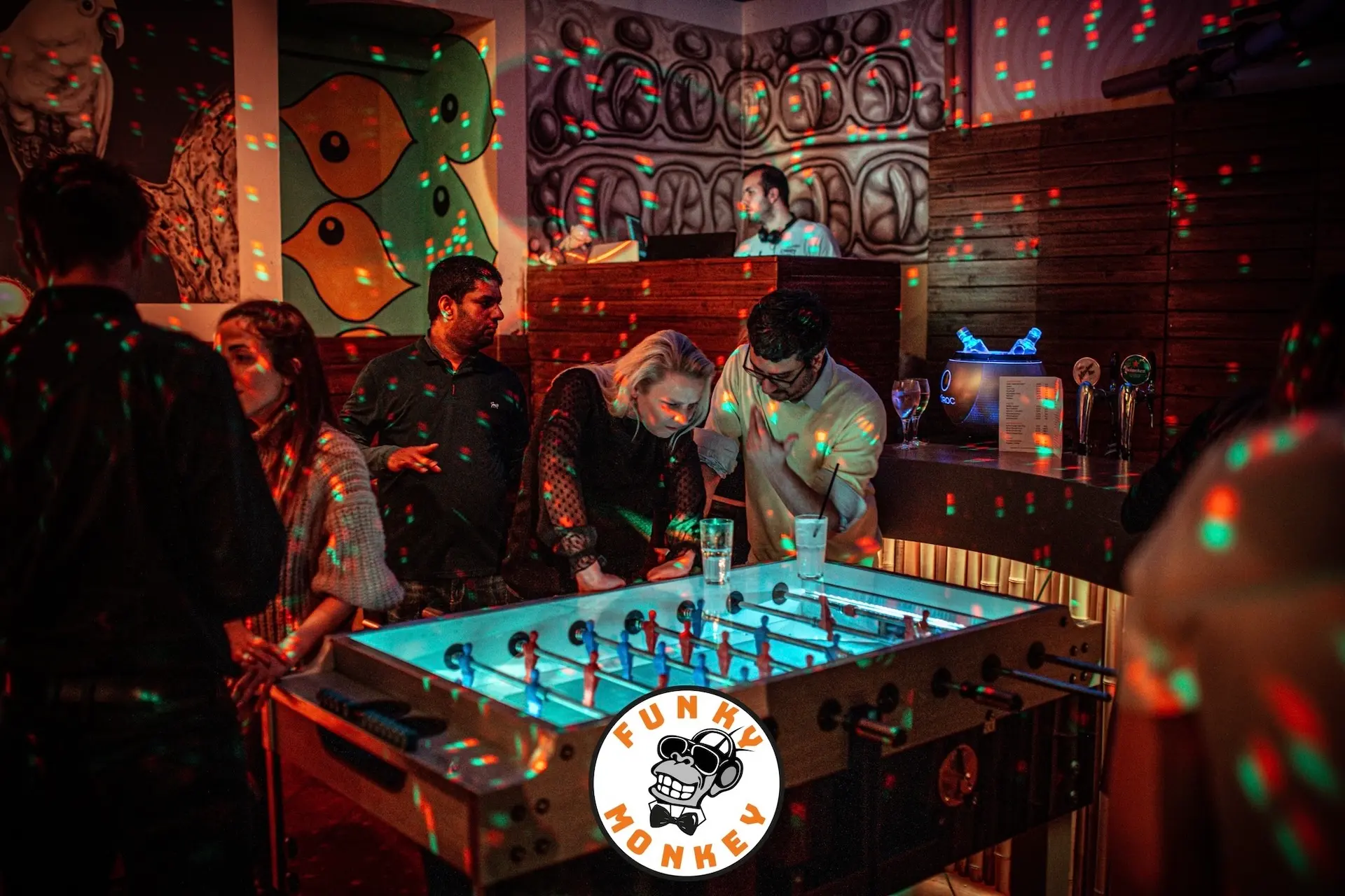 In einer lebhaften Bar mit moderner Kunst an den Wänden wird unter bunten Discolichtern Tischfußball gespielt. Im Vordergrund ist ein Funky Monkey-Logo zu sehen. Andere Gäste unterhalten sich und entspannen sich im Hintergrund.