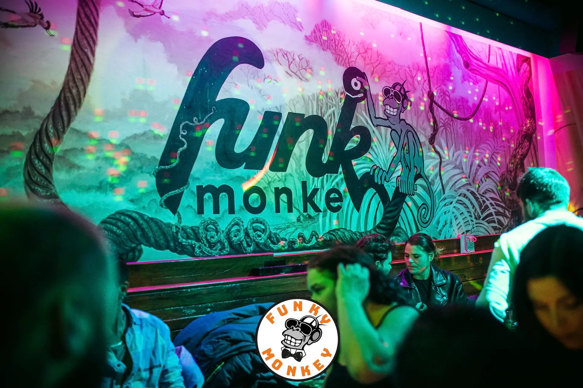 Bunte Nachtclubszene mit Menschen, die sich vor einer Wandmalerei mit dem Schriftzug "Funky Monkey" in fetten Lettern unterhalten, die einen Cartoon-Affen und lebhafte Dschungelkunstwerke zeigt. Die Beleuchtung ist lebhaft und farbenfroh.