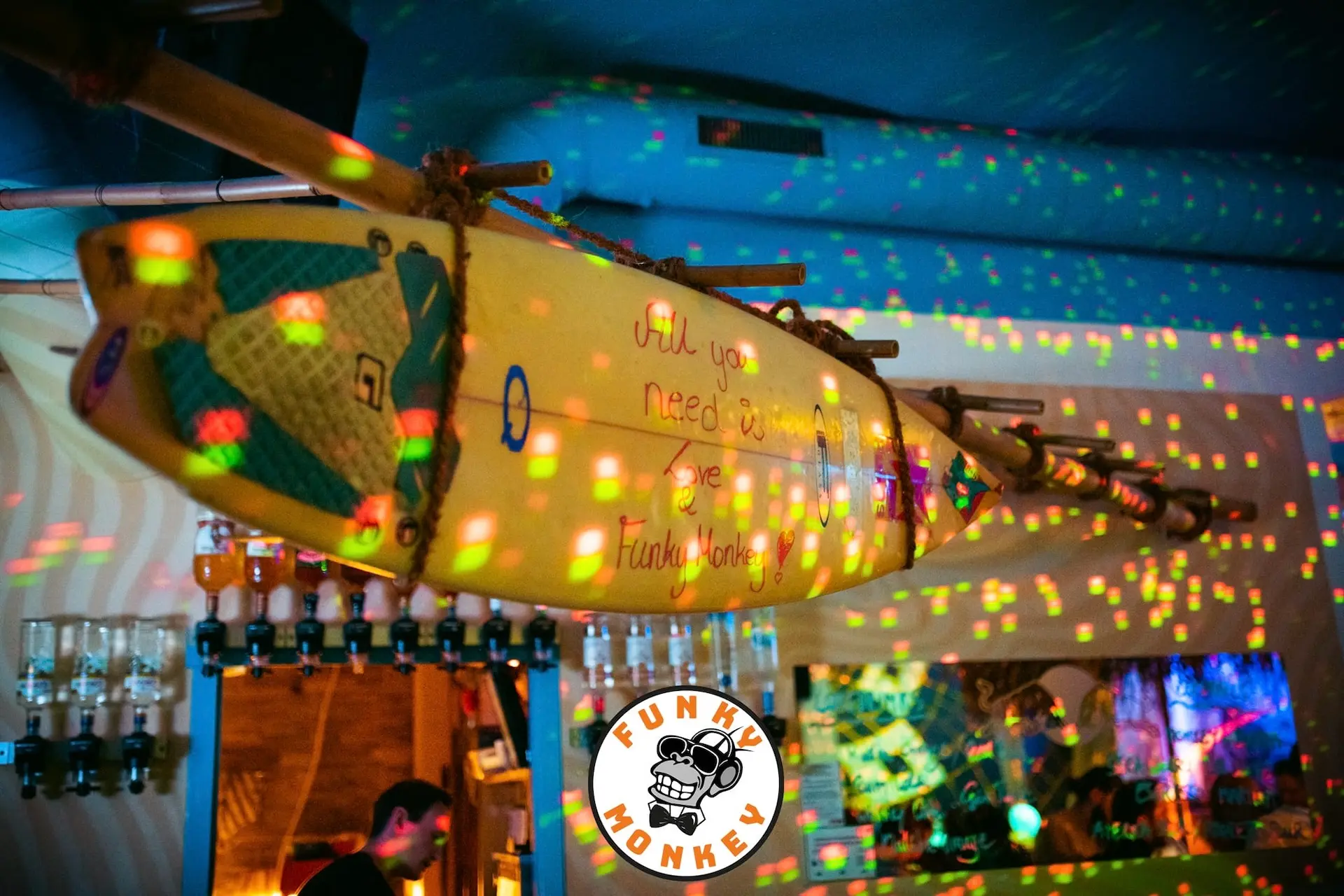 In einer farbenfrohen, neonbeleuchteten Bar hängt ein Surfbrett mit der Aufschrift "All you need is love" von der Decke, und am unteren Rand des Bildes ist das Funky-Monkey-Logo zu sehen.